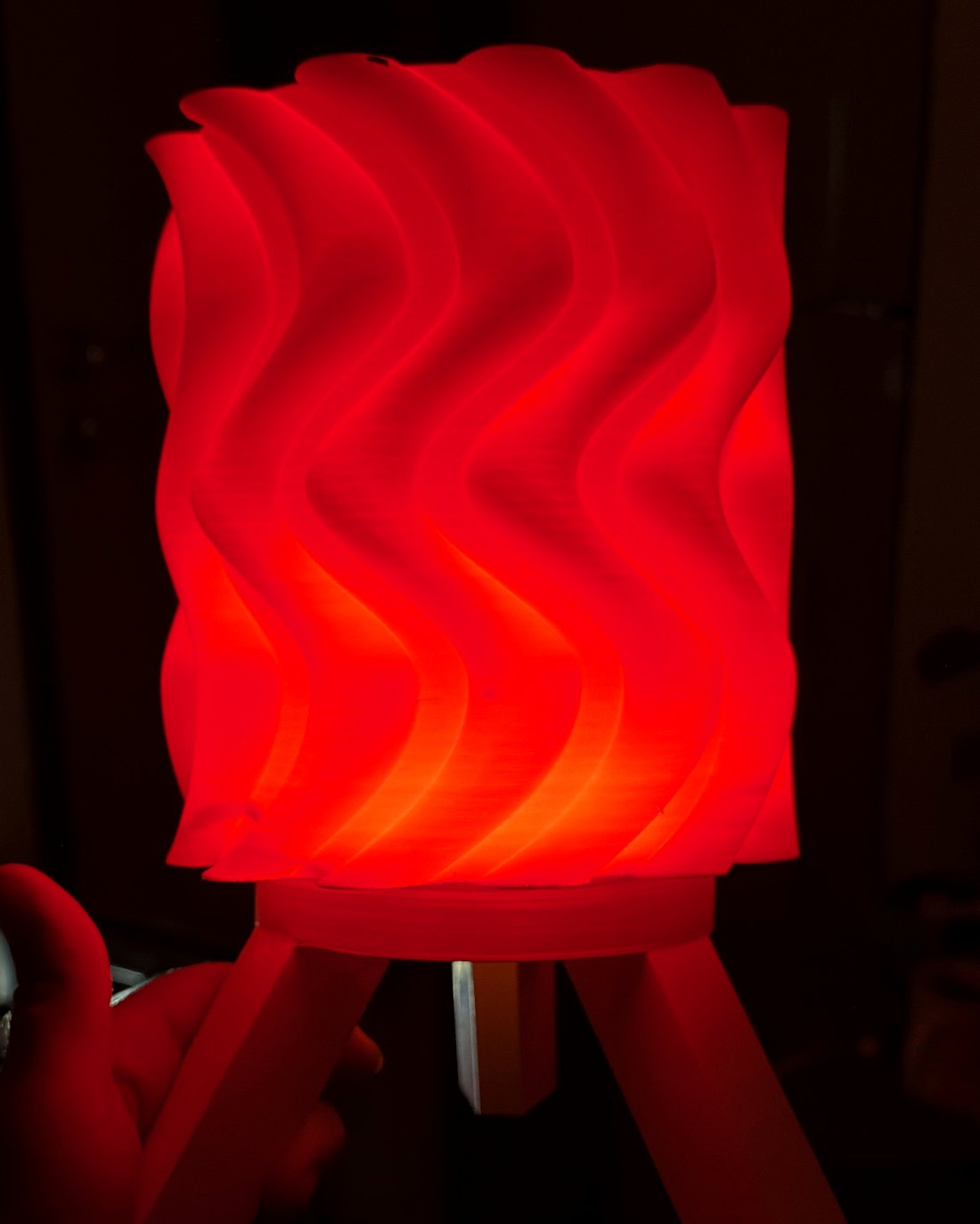Red 3D printed spiral wave lamp on tripod legs glowing warmly from within, dark studio product photo.